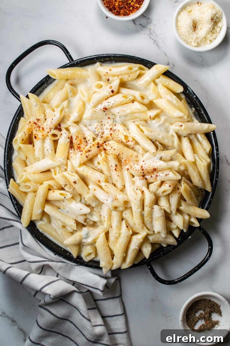 Top down view of a black pan of creamy pasta on a white backdrop with cheese and chili flakes in small white bowls.