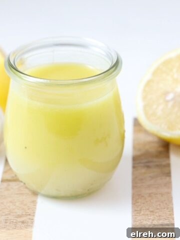 Lemon garlic salad dressing in a glass jar on a white and wooden board with fresh lemon halves and garlic.