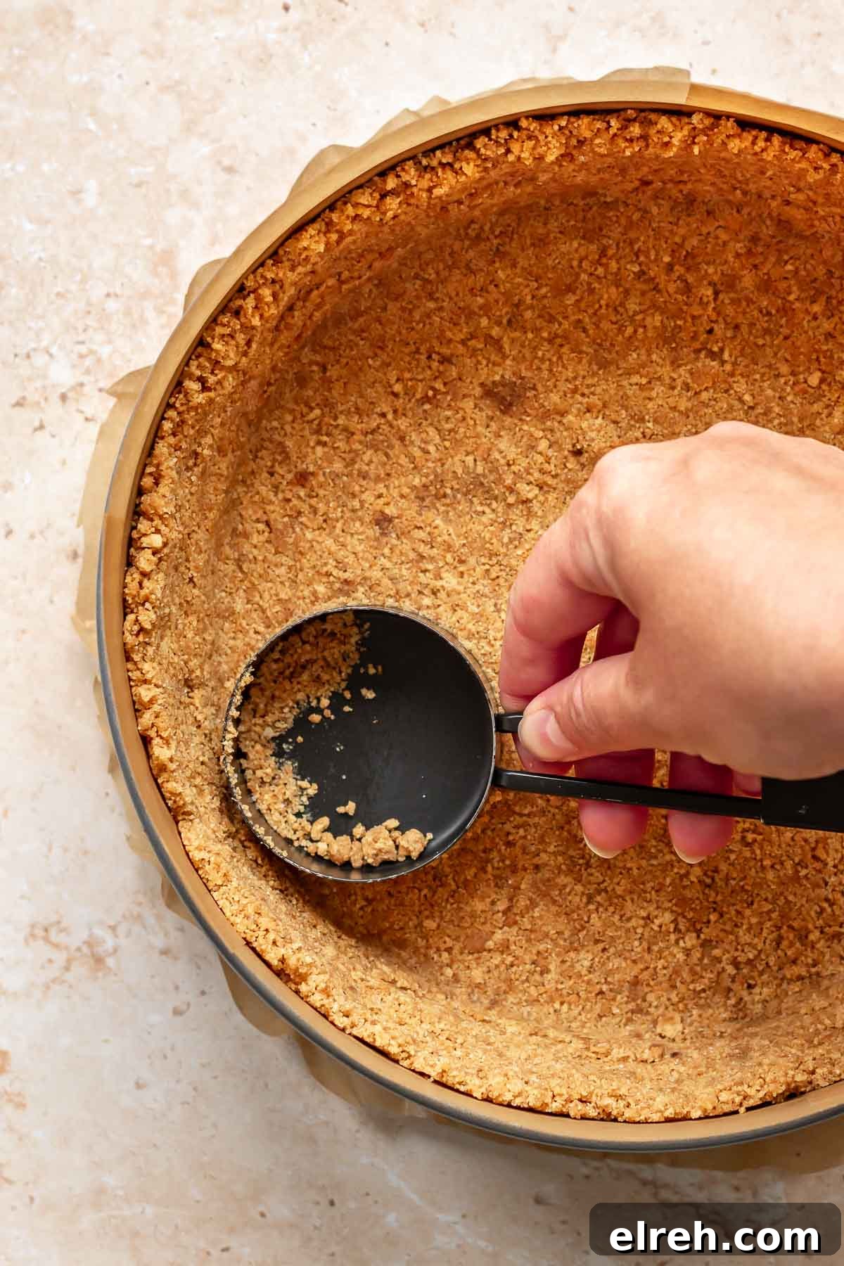 Apple Crumble Cheesecake with Cinnamon Streusel Recipe 9 A hand uses a measuring cup to press graham cracker crumbs into a springform pan.