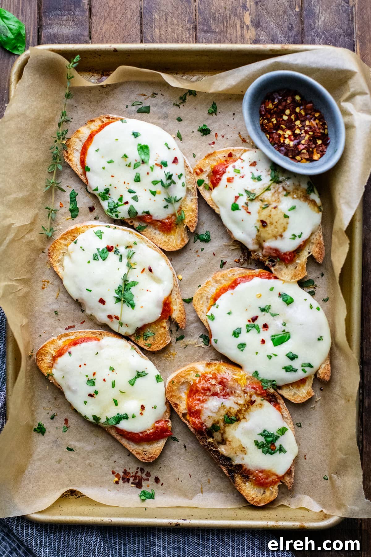 crostini pizzas arranged on baking sheet lined with parchment paper with fresh herbs on brown wooden background.