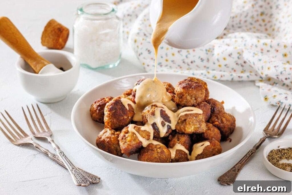A bowl of meatballs is topped with creamy sauce being poured from a jug, surrounded by forks and seasoning jars on a white table.