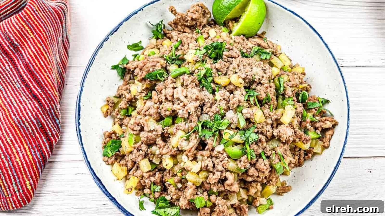 A plate of cooked ground beef mixed with diced vegetables and garnished with lime wedges and chopped herbs. A red striped cloth is placed beside the plate.