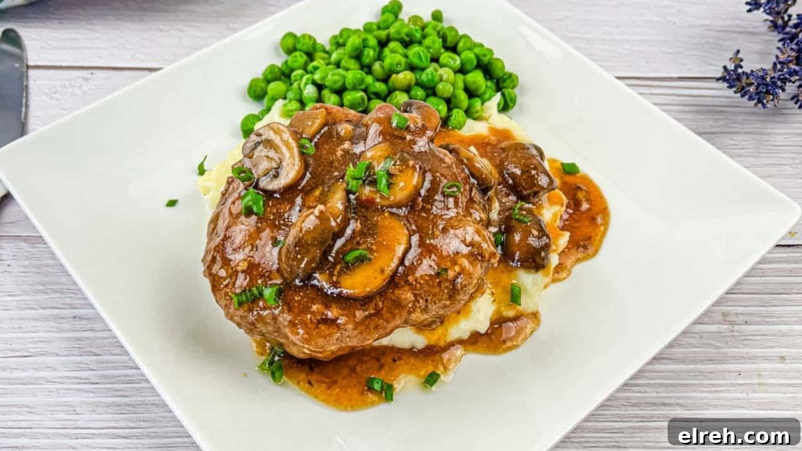 A white plate with Salisbury steak topped with mushroom gravy on mashed potatoes, accompanied by a side of green peas.