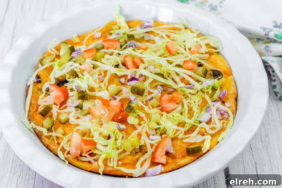 A Cheeseburger Pie topped with shredded lettuce, diced tomatoes, pickles, and chopped red onions in a white pie dish on a light wooden surface.