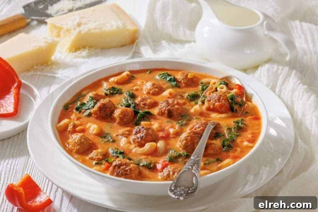 A bowl of meatball soup with pasta, spinach, and tomato broth, served with a spoon.