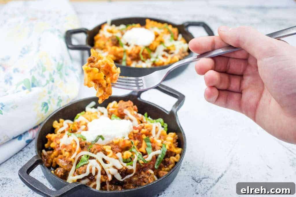 A hand holds a fork with a Classic Skillet Lasagna with Ground Beef topped with melted cheese and herbs.