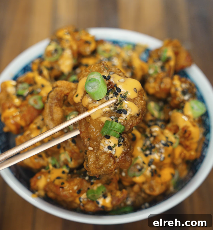 How to Make Mochiko Chicken: Crispy Hawaiian Fried Chicken 3 A crispy, golden mochiko chicken bite held with chopsticks above a bowl, showing off garnishes of scallions and sesame seeds.