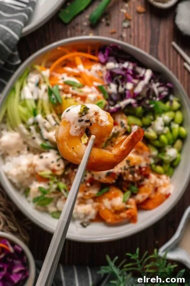 A close-up of chopsticks holding a cooked shrimp above a bowl filled with rice, shredded vegetables, edamame, and sauce.