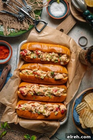 Four split top toasted buns filled with creamy lobster salad, garnished with chopped vegetables, are arranged on parchment paper next to chips and seasonings on a rustic table.