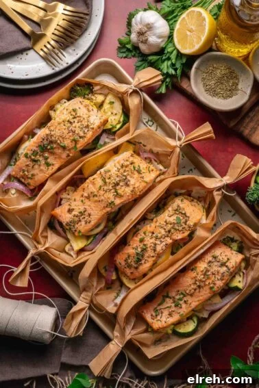 Four salmon fillets seasoned with herbs are placed on top of mixed vegetables and wrapped in parchment paper boats, displayed on a baking tray with utensils and ingredients nearby.