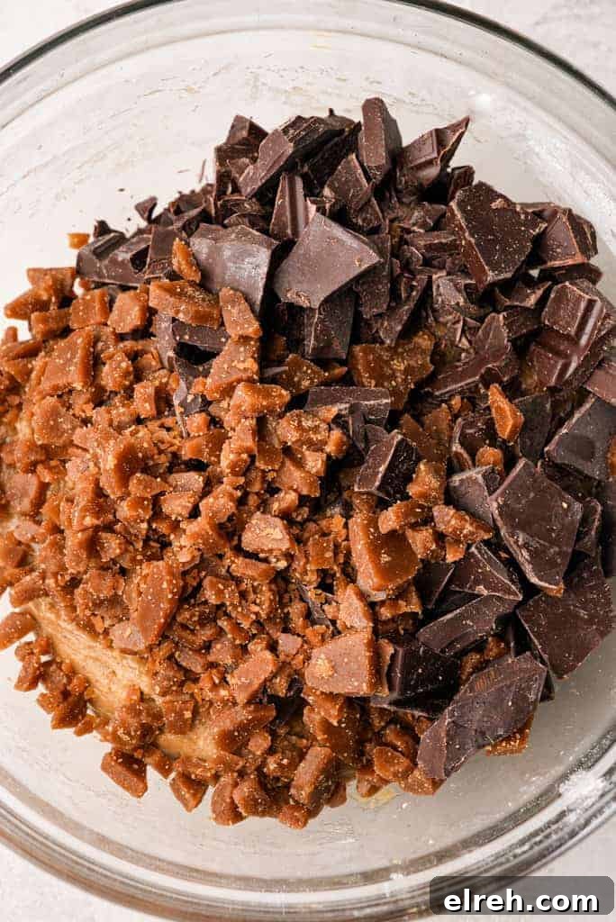 A glass mixing bowl filled with cookie dough topped with chopped chocolate chunks and crushed toffee pieces.