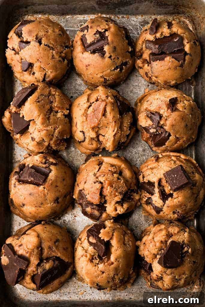 Twelve rounded scoops of brown butter toffee chocolate chip cookie dough studded with large chunks of chocolate are arranged in rows on a metal baking sheet.