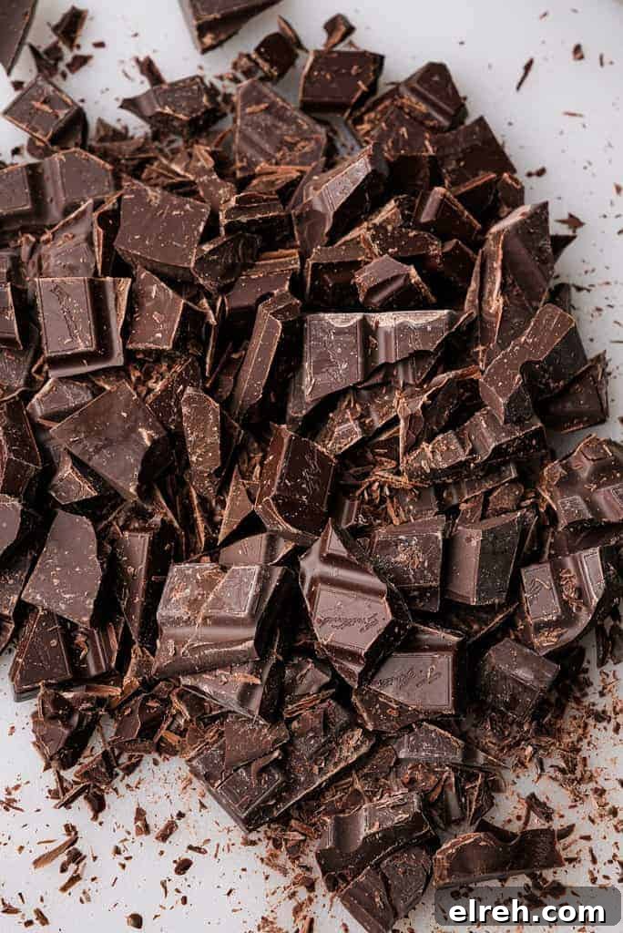 A close-up of roughly chopped dark chocolate pieces scattered across a white surface with chocolate shavings around them.