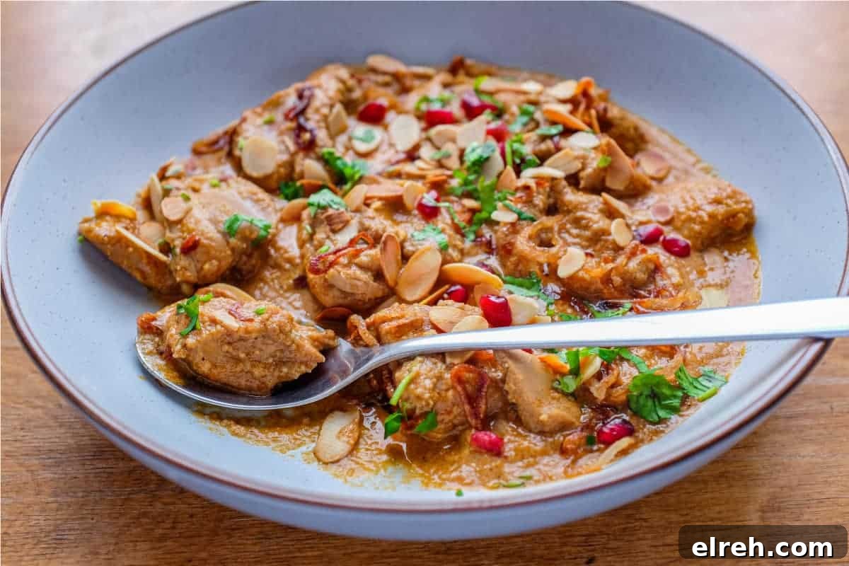 A bowl of Mughlai Chicken Curry garnished with Almonds, cilantro and pomegranate seeds with a spoon on the side.