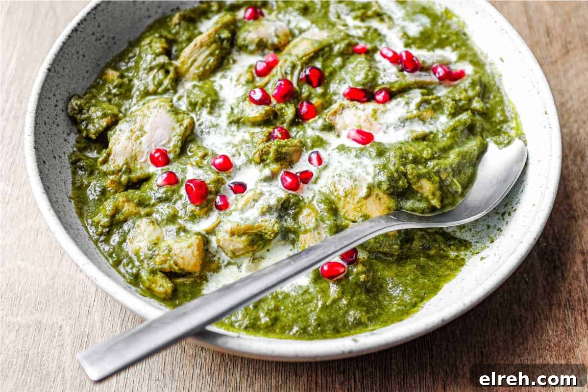 A bowl of Chicken Saag curry with a spoon, drizzled with fresh cream and sprinkled with pomegranate seeds.