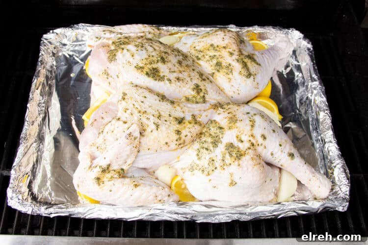 Angled, overhead view of a spatchcocked turkey covered in compound butter resting in an aluminum disposable pan lined with foil on a grill grate.