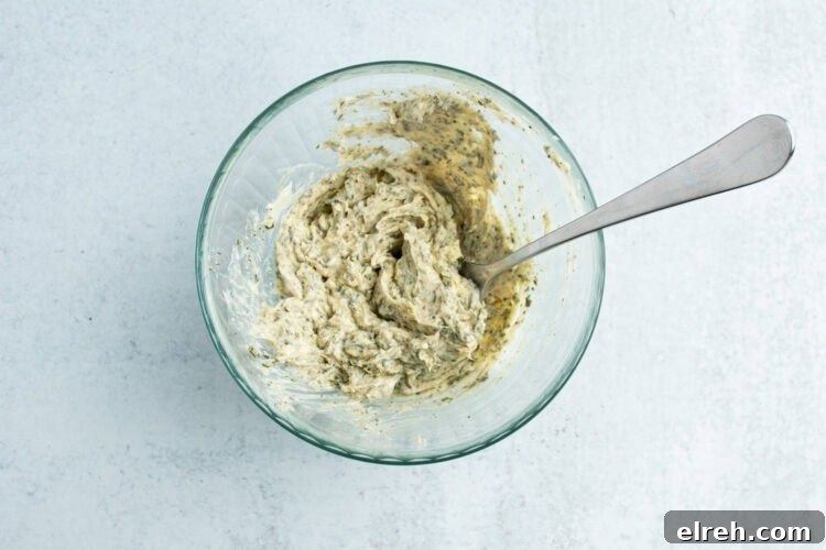 Overhead view of a small glass mixing bowl holding a compound butter mixture of garlic and fresh herbs.
