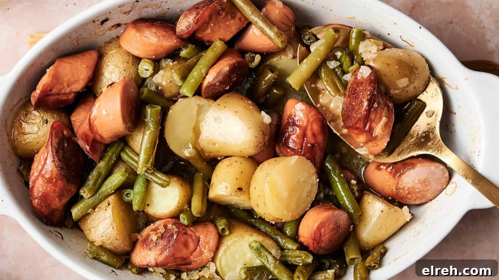 A baked dish containing sliced sausages, potatoes, and green beans in a white casserole dish with a serving spoon.