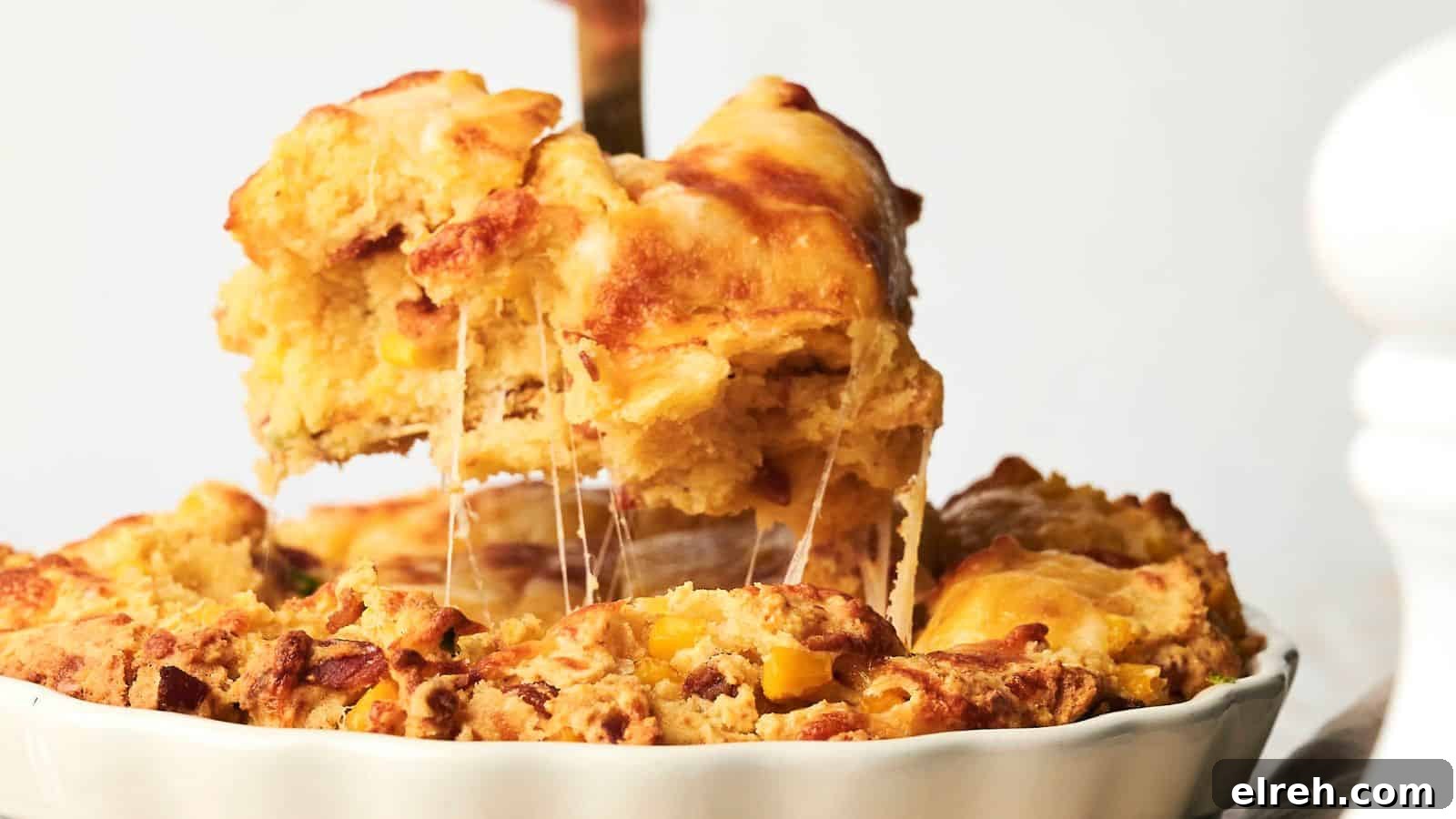 A portion of a cheesy, golden-brown savory cornbread casserole is being lifted from the dish, showing its flaky crust and gooey cheese filling.