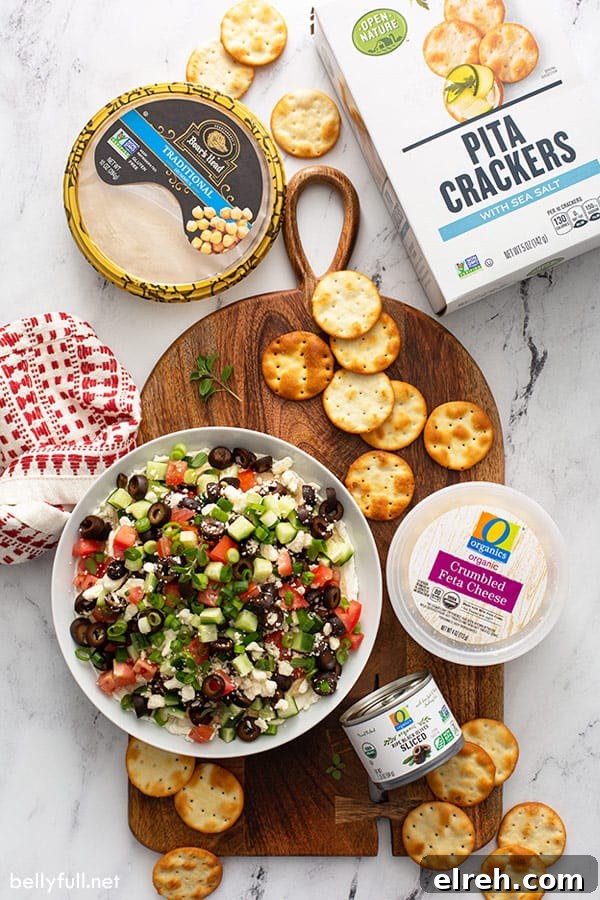 layered dip with cream cheese, hummus, olives, cucumber, and tomato