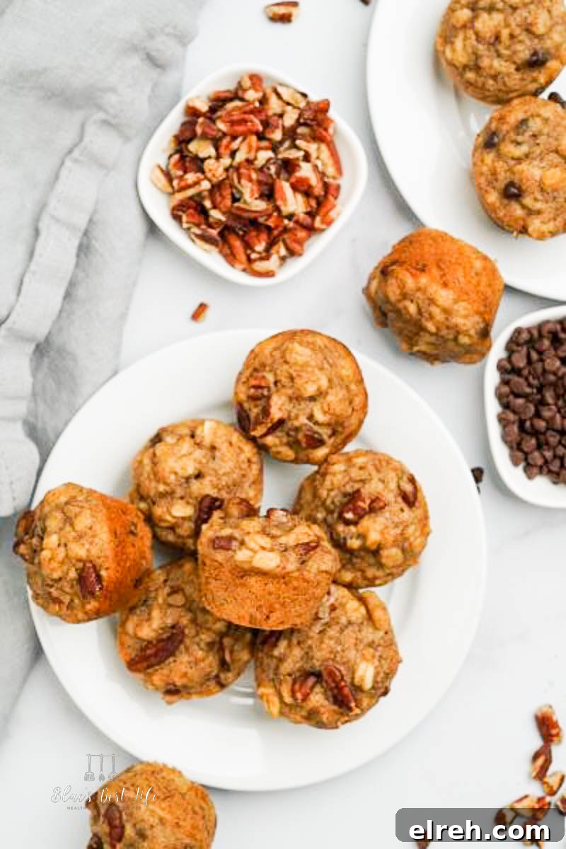 Top-down view of freshly baked banana muffins with pecans and chocolate chips on white plates; surrounded by small bowls of extra ingredients and a gray cloth napkin.