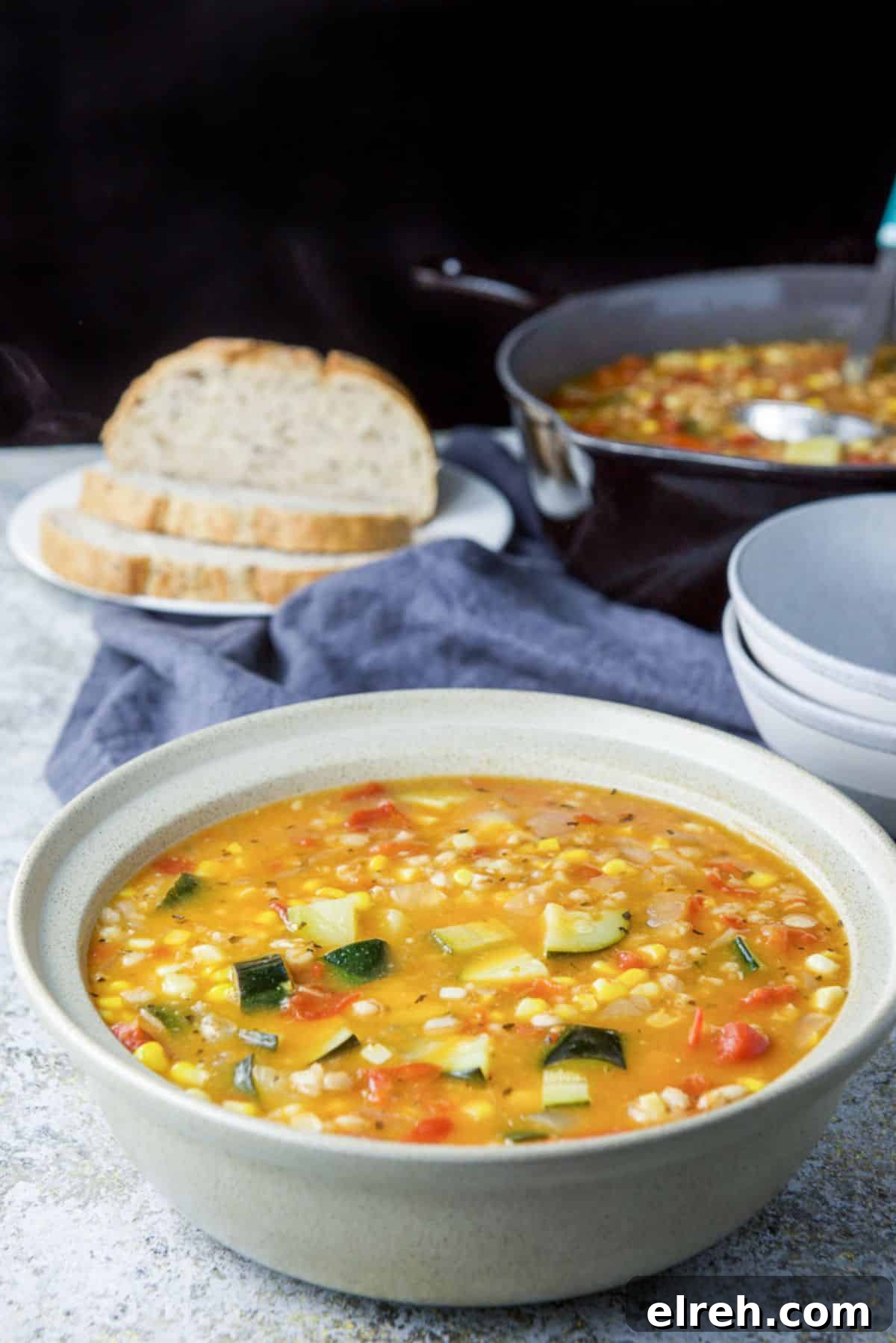 Soup with corn and zucchini in a serving bowl with the bread, a pan and bowls in the background