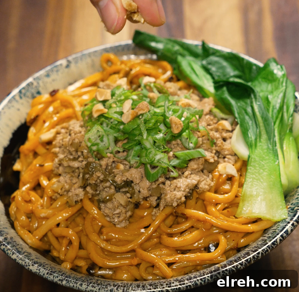 Hand sprinkling crushed peanuts onto a bowl of dan dan noodles topped with pork, scallions, and bok choy.