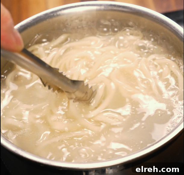 Noodles boiling in a pot of water being stirred with tongs — prepping the base for dan dan noodles.