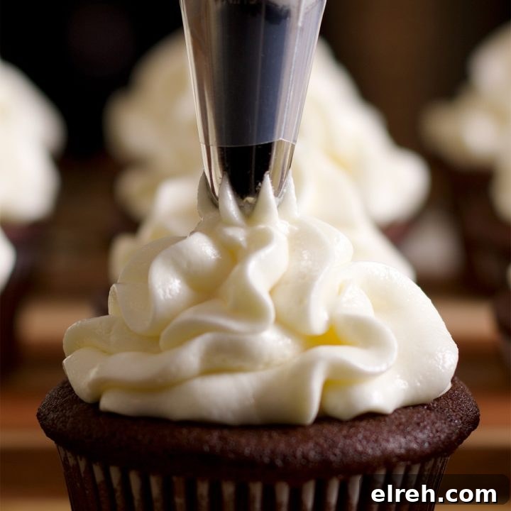 Someone piping cream cheese buttercream from a pastry bag onto the top of a chocolate cupcake.