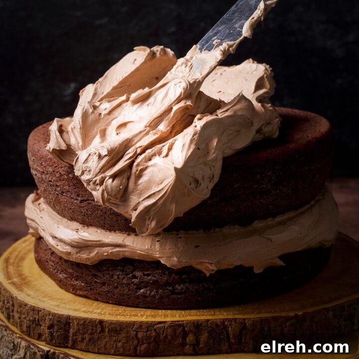 Someone using an offset icing spatula to spread milk chocolate buttercream over the top of a two-layer dark chocolate cake.