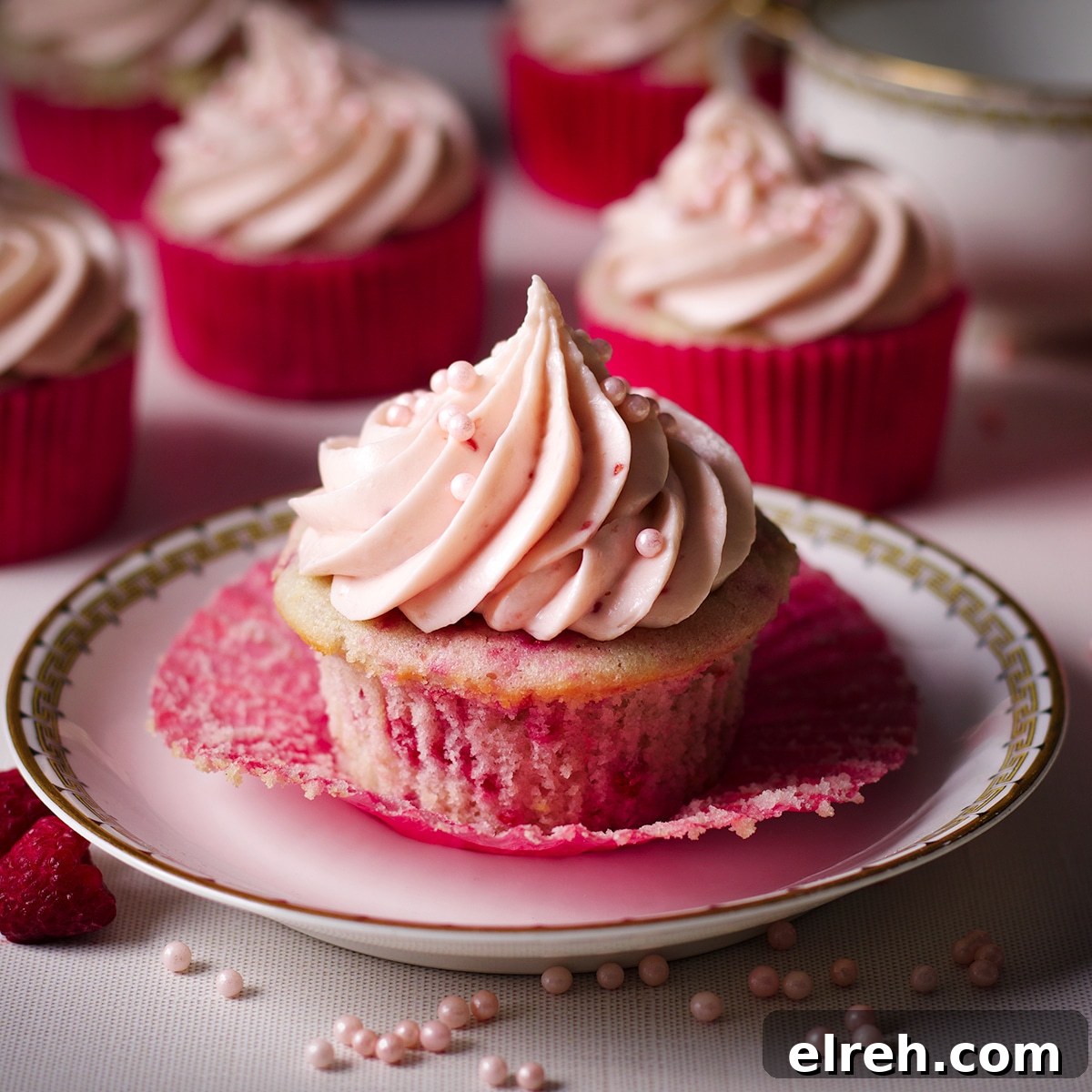 Raspberry Cupcakes Recipe with Cream Cheese Frosting 9 A raspberry cupcake on a dessert plate with several other cupcakes resting on the table behind it.