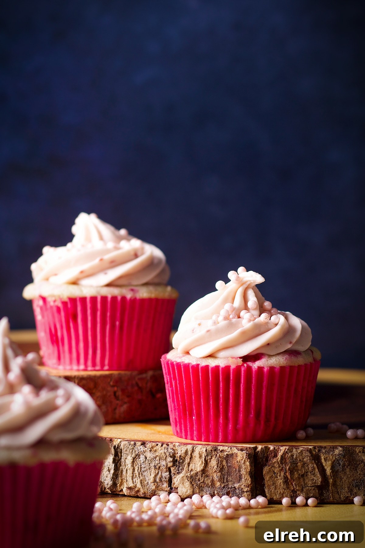 Raspberry Cupcakes Recipe with Cream Cheese Frosting 10 Three raspberry cupcakes topped with a swirl of raspberry cream cheese buttercream on a wood cake stand.
