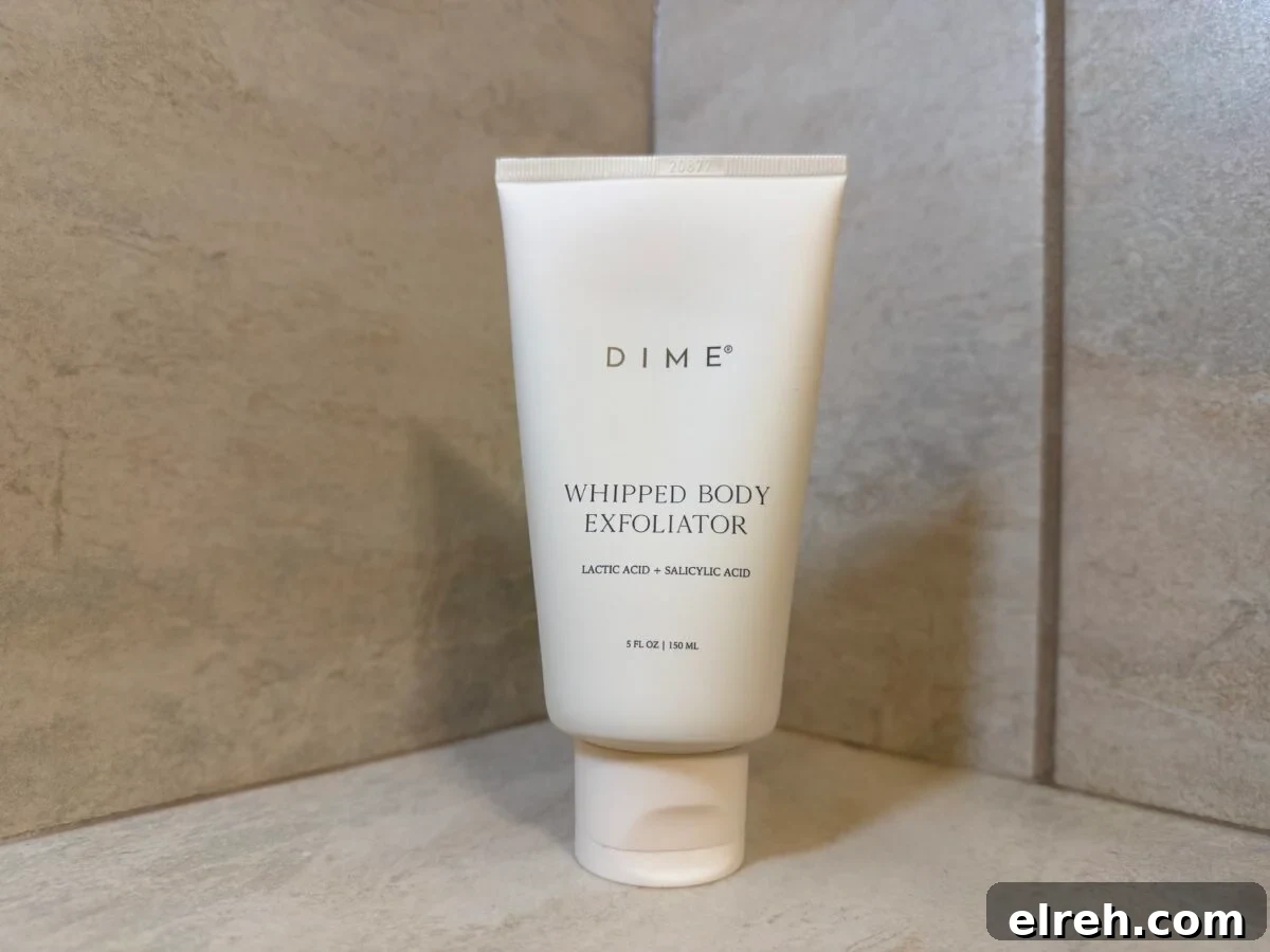 Tube of Dime Whipped Body Exfoliator standing on a tiled shower shelf.