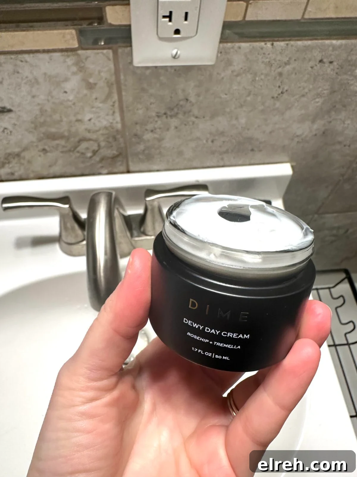 A hand is holding an open jar of DIME Dewy Day Cream