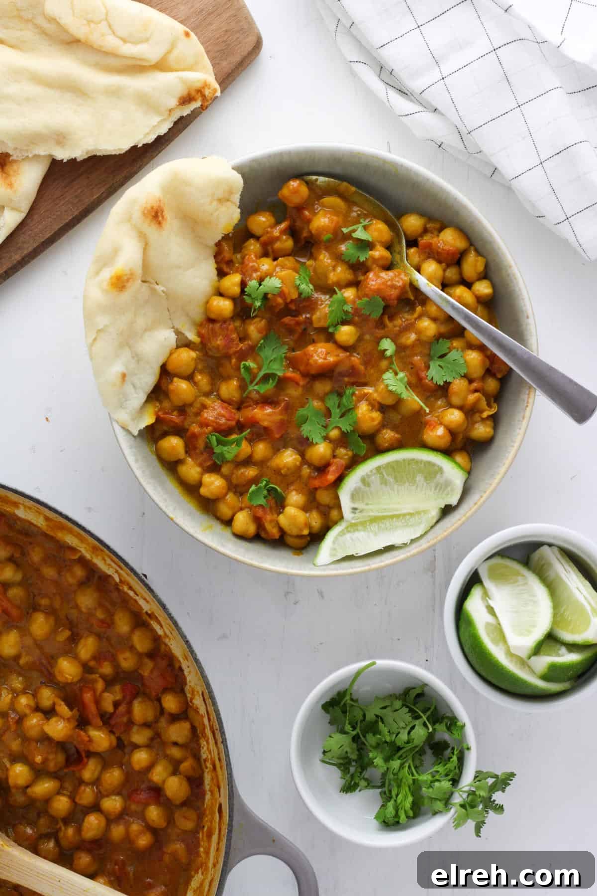 A white bowl with golden brown coconut chickpea curry, a spoon, a couple wedges of lime and a torn piece of naan. Just off center is more naan, lime wedges and more curry.