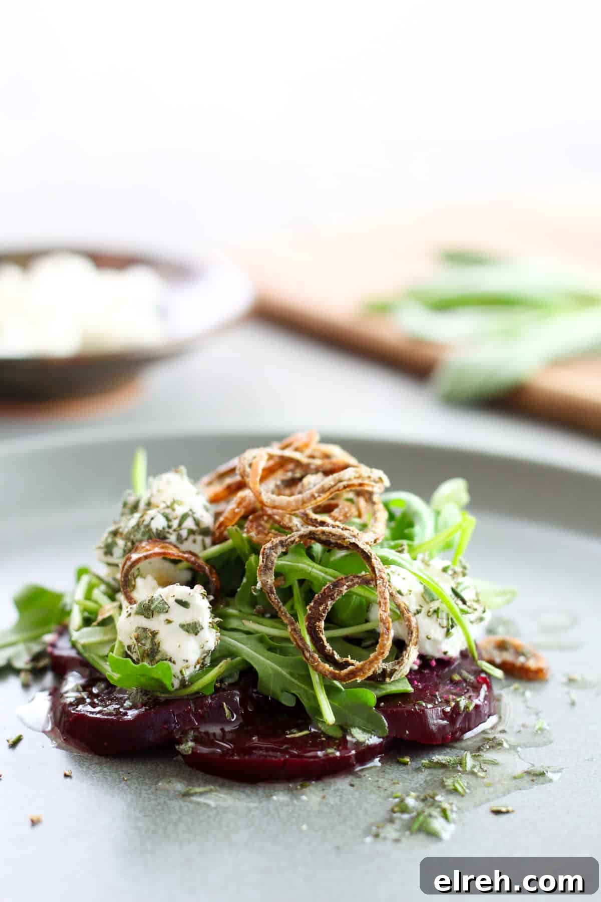 A close up of sliced purple beets topped with arugula, goat cheese rolled in fresh herbs, and topped with crispy fried shallots all on a gray plate. Out of focus in the background is more goat cheese on a small bowl and herbs on a cutting board.