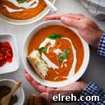 A bowl of lentil and red pepper soup held by two hands with another bowl of soup off center. The soup is topped with yogurt, basil and bread.
