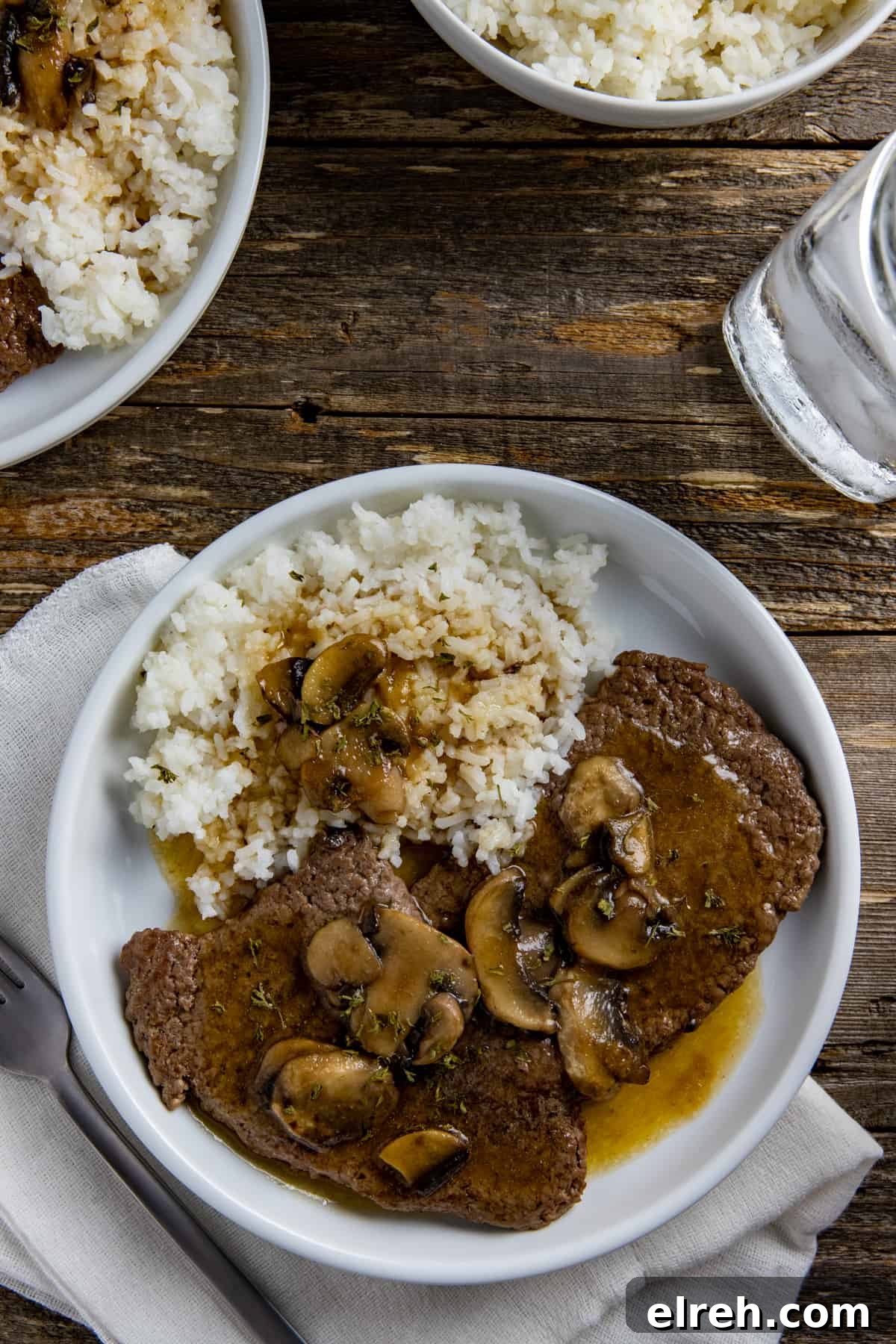 Two cooked steaks covered with gravy and mushrooms with a side of rice and gravy.