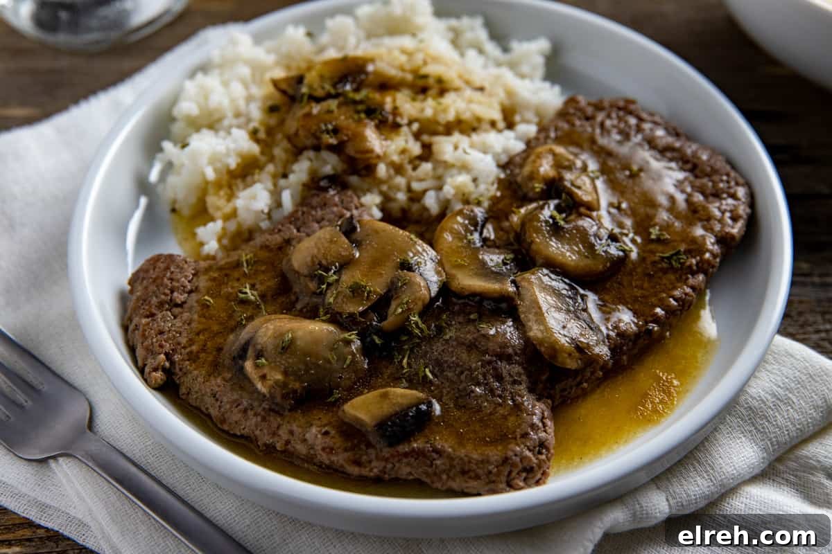 Two cooked steaks covered with gravy and mushrooms with a side of rice and gravy.