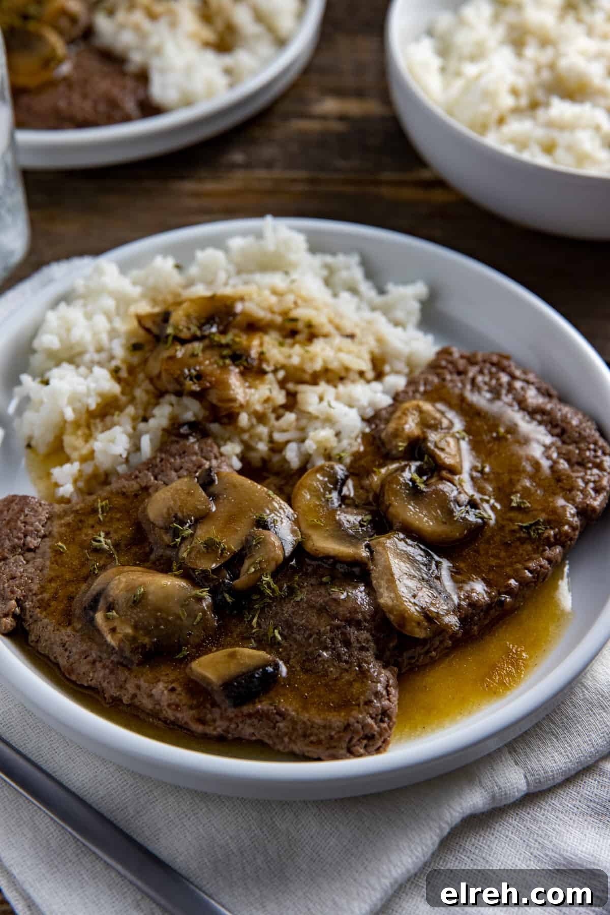 Two cooked steaks covered with gravy and mushrooms with a side of rice and gravy.
