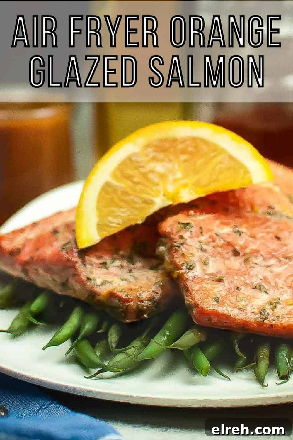 Side shot of two salmon filets garnished with an orange slice on a bed of green beans on a square white plate with a blue cloth and fork on the side.
