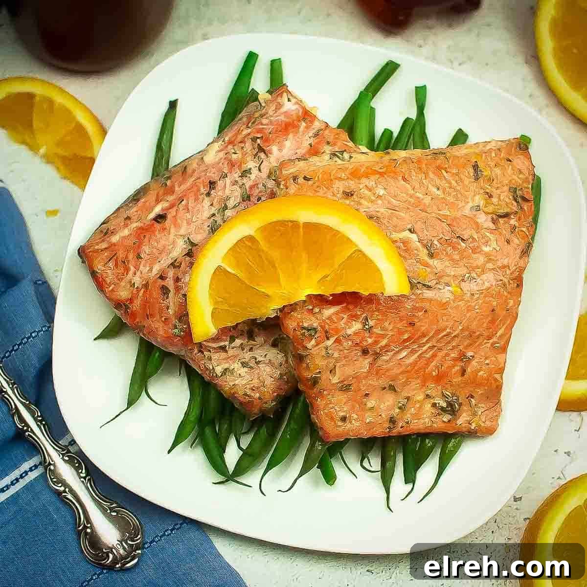 Overhead shot of two salmon filets garnished with an orange on a bed of green beans on a white square plate.