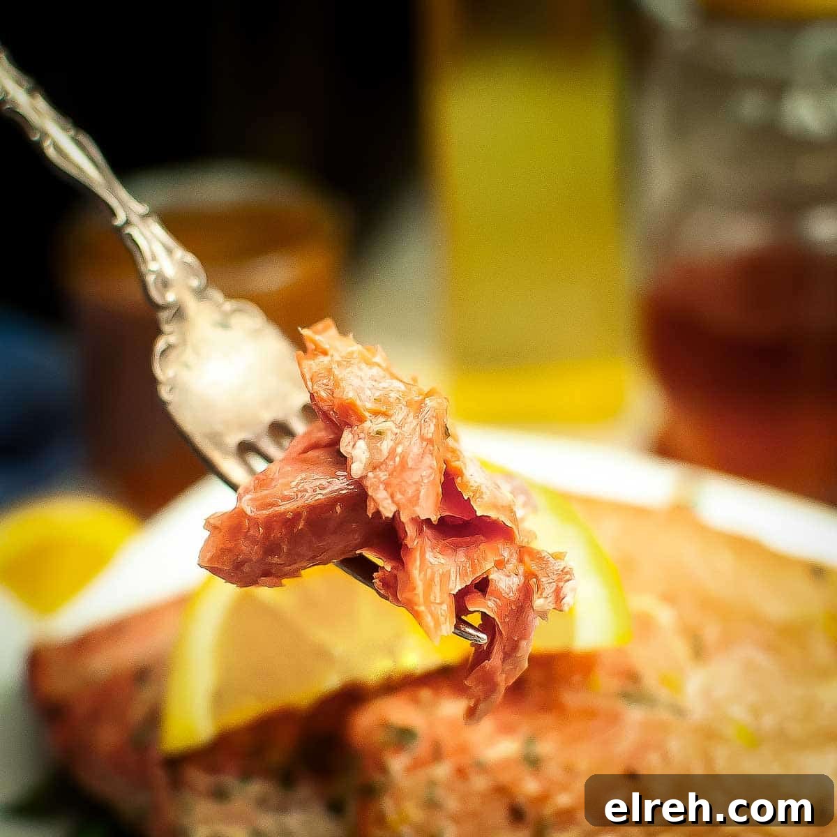 Side close up shot of a fork holding up a piece of salmon over a white plate with two salmon filets garnished with an orange slice.