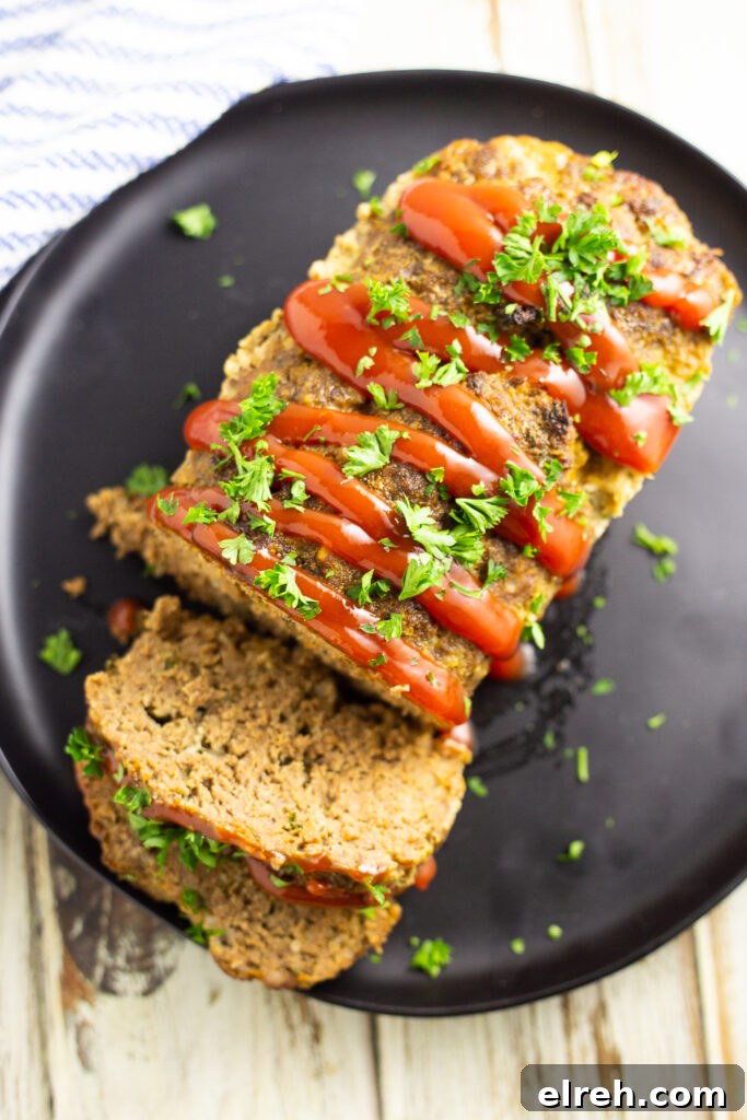 Overhead view of a full meatloaf on a large plate with 2 slices cut off garnished with ketchup drizzle and fresh chopped parsley.