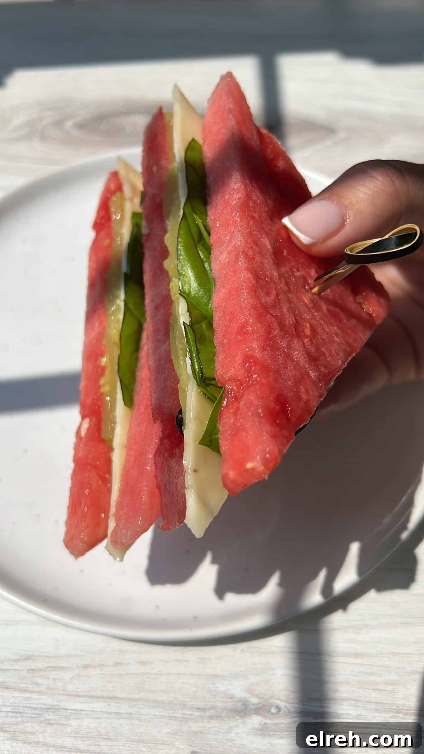 Watermelon Sandwich Recipe for a Refreshing Summer Snack 2 A hand holds a sandwich made with watermelon slices as the bread, filled with cheese and leafy greens, on a white plate. Bright natural light casts shadows across the scene.
