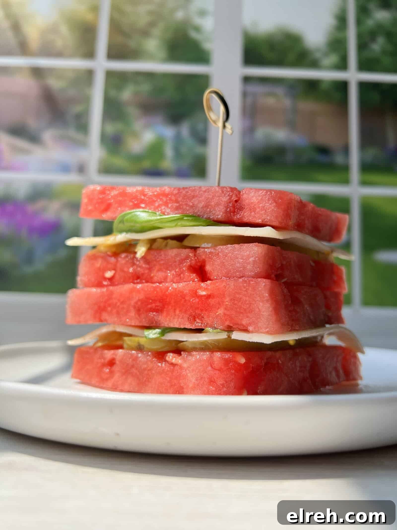 Watermelon Sandwich Recipe for a Refreshing Summer Snack 3 A stack of watermelon slices layered with mozzarella cheese and basil, secured with a wooden pick, sits on a white plate in front of a sunlit window with a garden view.