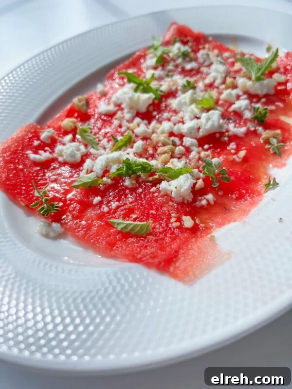 Thin slices of watermelon carpaccio arranged on a white plate with crumbled feta and herbs.