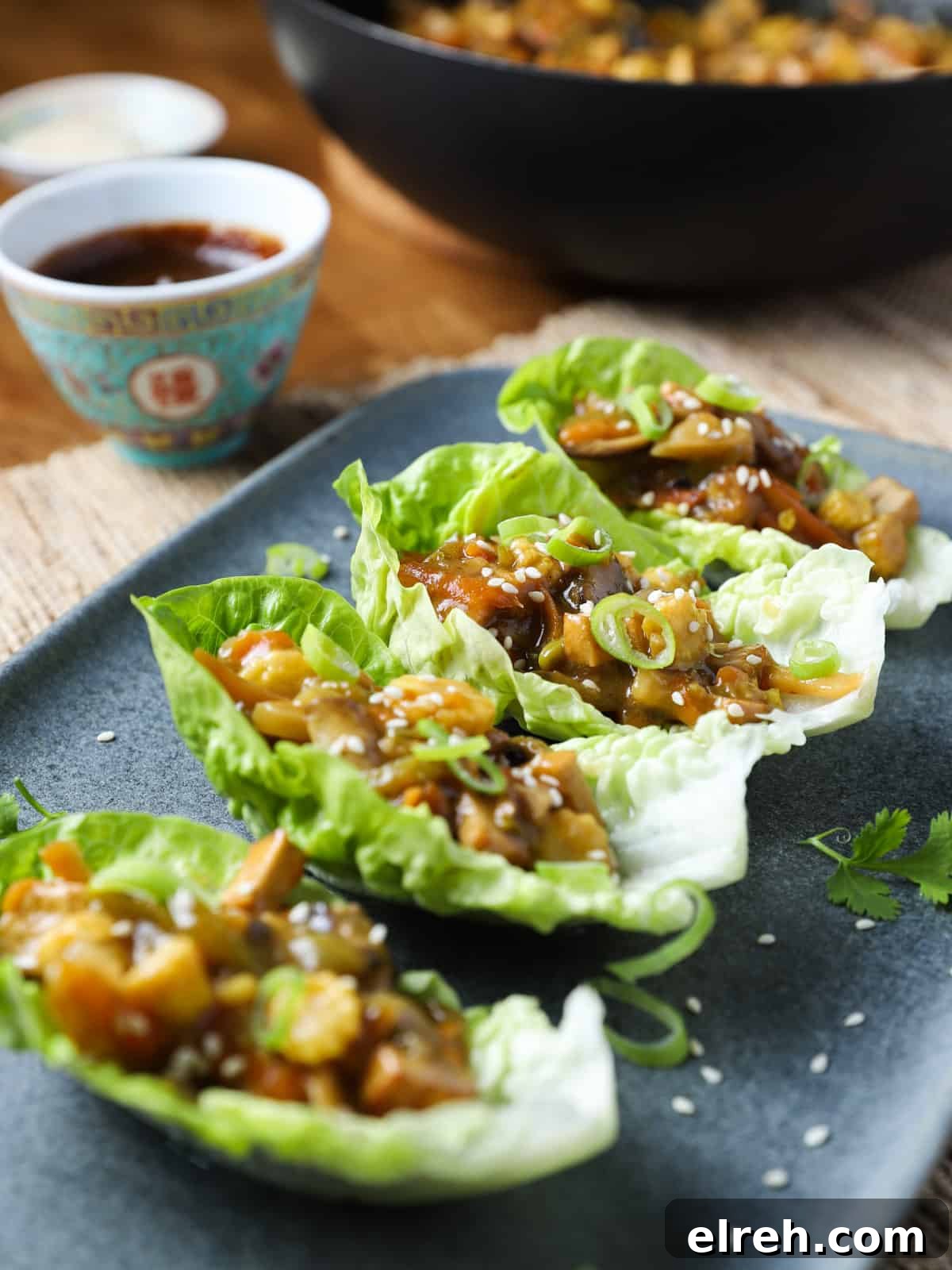 Vegan San Choy Bau Lettuce Cups 2 Photo of 4 lettuce leaves with vegan san choy bau filling inside, placed on a blue-grey oblong plate with pan in the background and a chinese dish of hoisin sauce to the side.