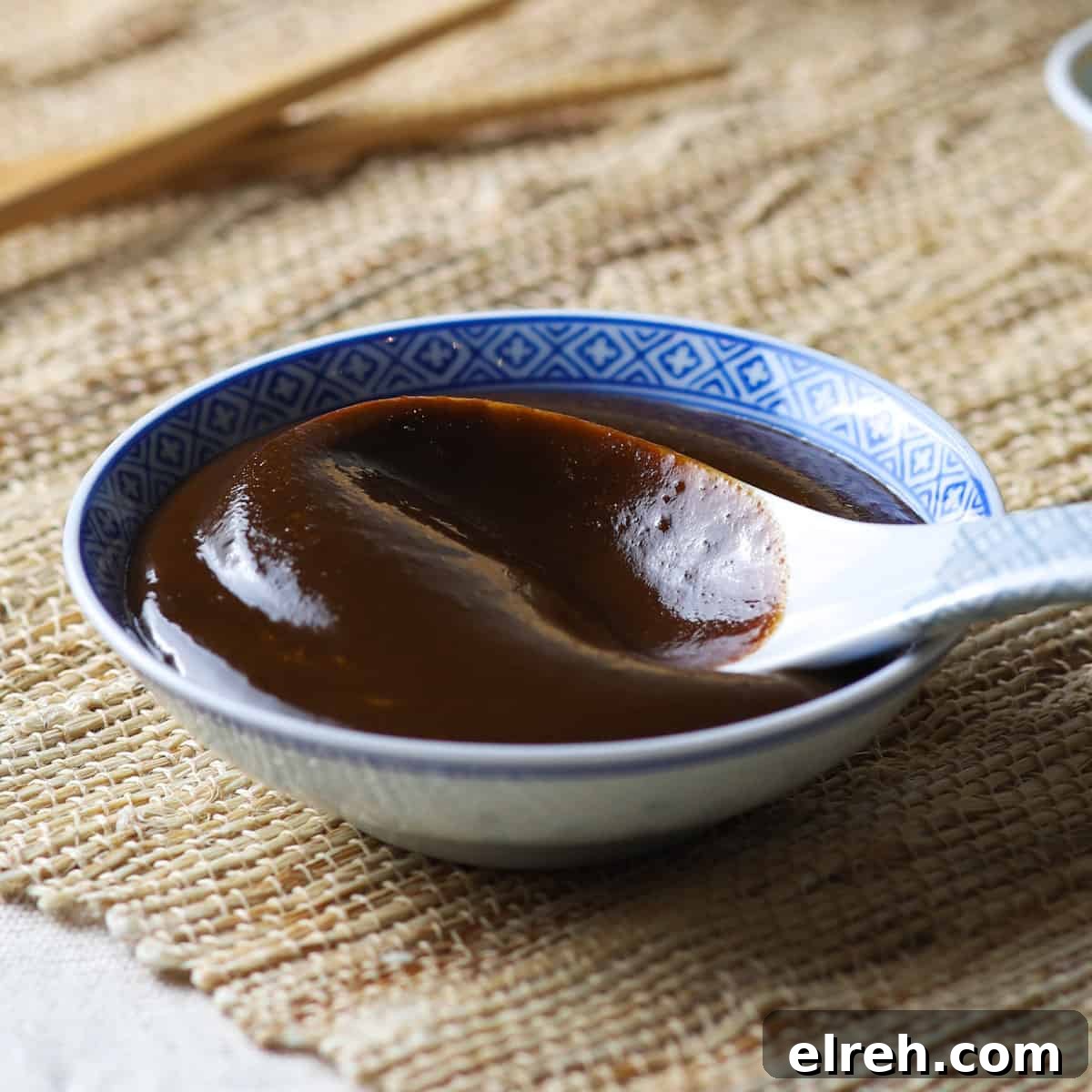 Close-up photo of a shallow Chinese bowl filled with homemade hoisin sauce and a Chinese ceramic spoon, all on a jute mat with bamboo steamer and chop sticks in the background.
