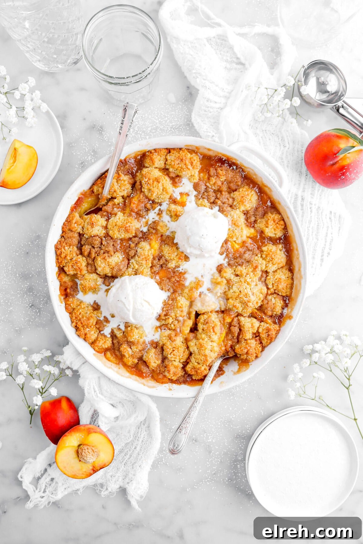 Peach Cobbler Crisp with Brown Sugar and Cinnamon 19 peach cobbler with spoons dug into it from the top and bottom of casserole with two scoops of melting ice cream on top, peaches and white flowers around on marble surface.